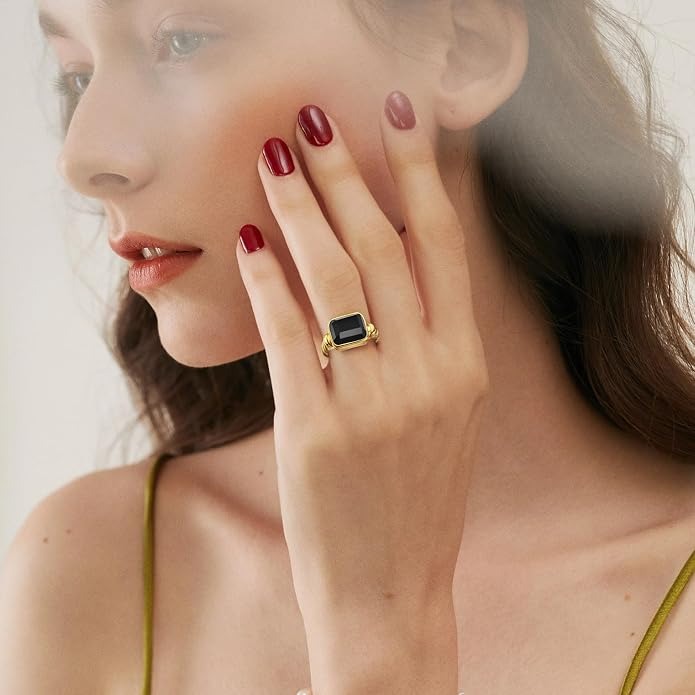 A close-up lifestyle shot of a model wearing a gold ring with a rectangular dark blue stone, with her hand resting on her cheek.