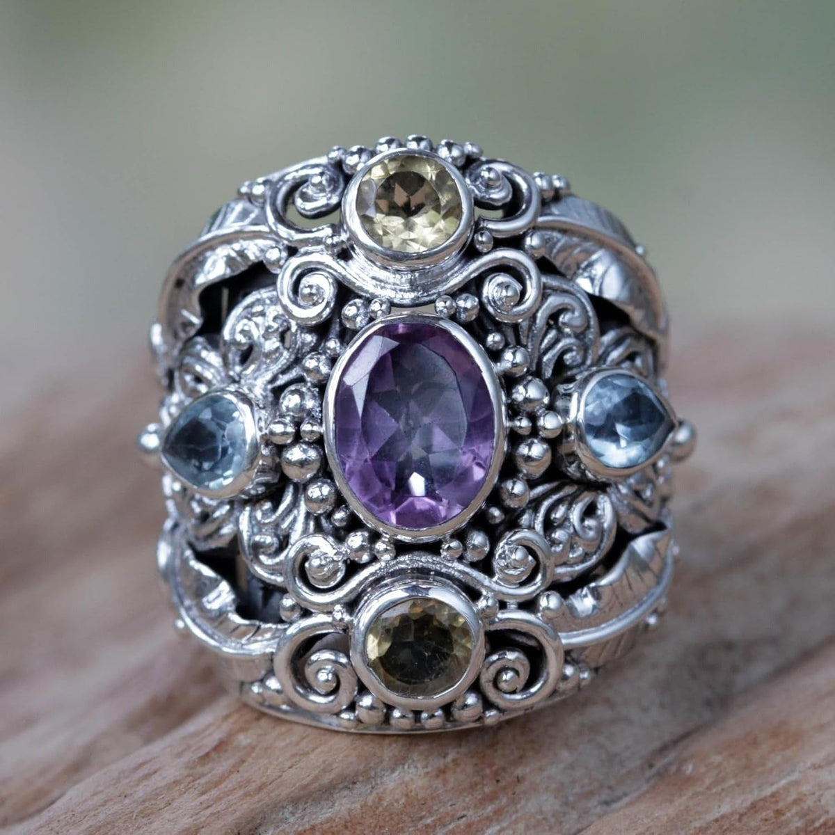 An ornate silver ring with a wide filigree band, featuring a large oval purple amethyst center stone and smaller round and teardrop-cut gemstones in yellow, blue, and green, resting on a wooden surface.