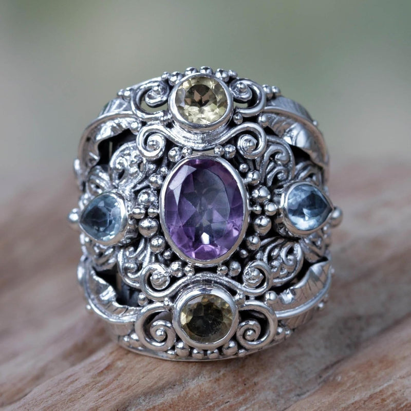 An ornate silver ring with a wide filigree band, featuring a large oval purple amethyst center stone and smaller round and teardrop-cut gemstones in yellow, blue, and green, resting on a wooden surface.