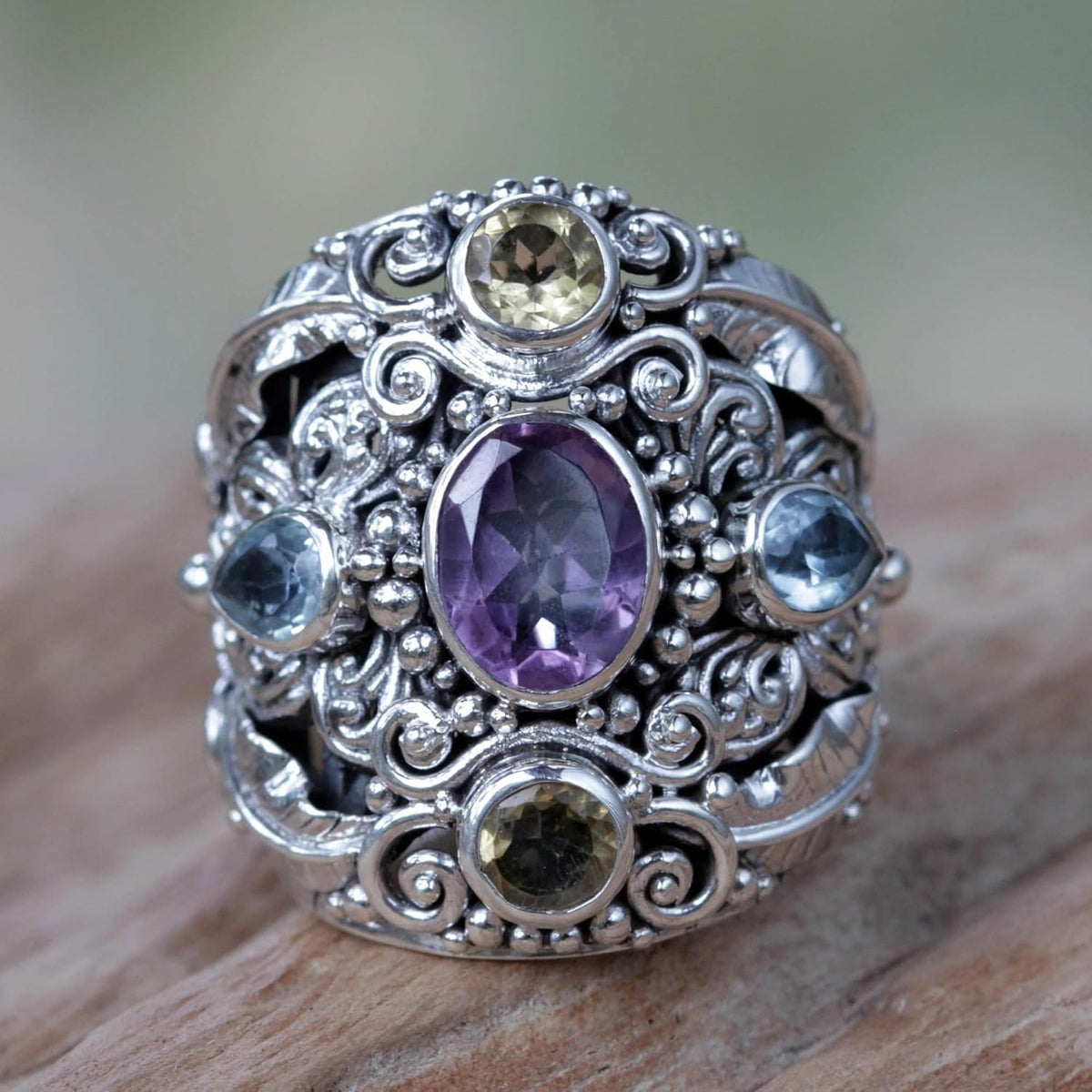 An ornate silver ring with a wide filigree band, featuring a large oval purple amethyst center stone and smaller round and teardrop-cut gemstones in yellow, blue, and green, resting on a wooden surface.
