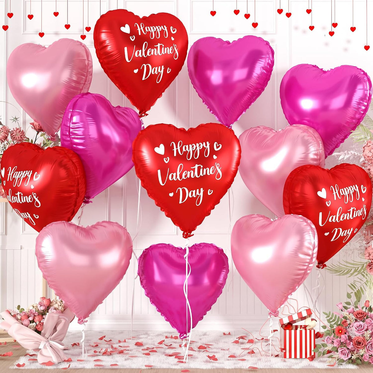 A set of metallic heart-shaped foil balloons in shades of red, pink, and light pink, decorated with 'Happy Valentine's Day' text, arranged with floral decorations and a gift box on a white paneled background.