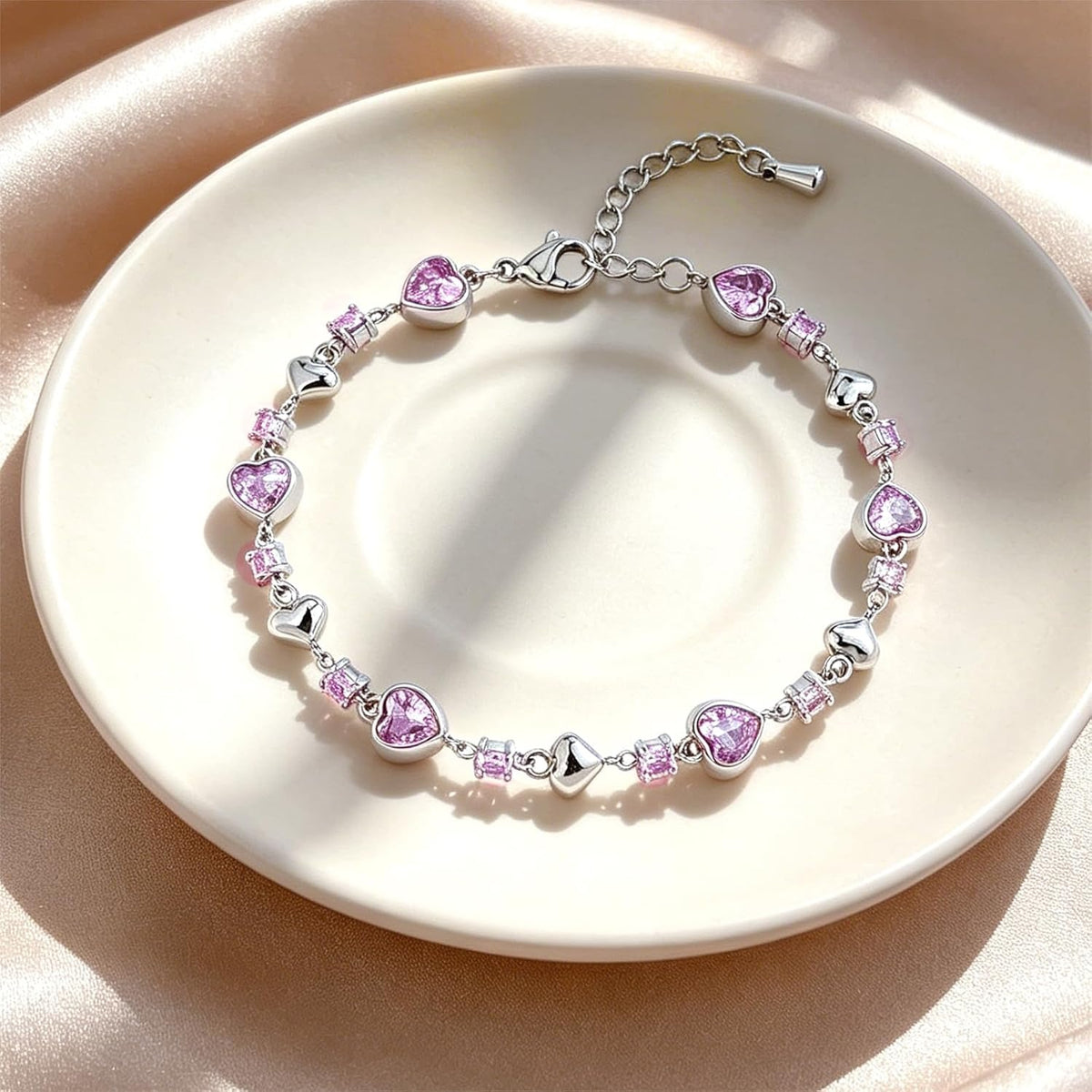 A silver bracelet with heart-shaped pink gemstones and small square pink gemstones, displayed on a round cream-colored plate against a satin fabric background.