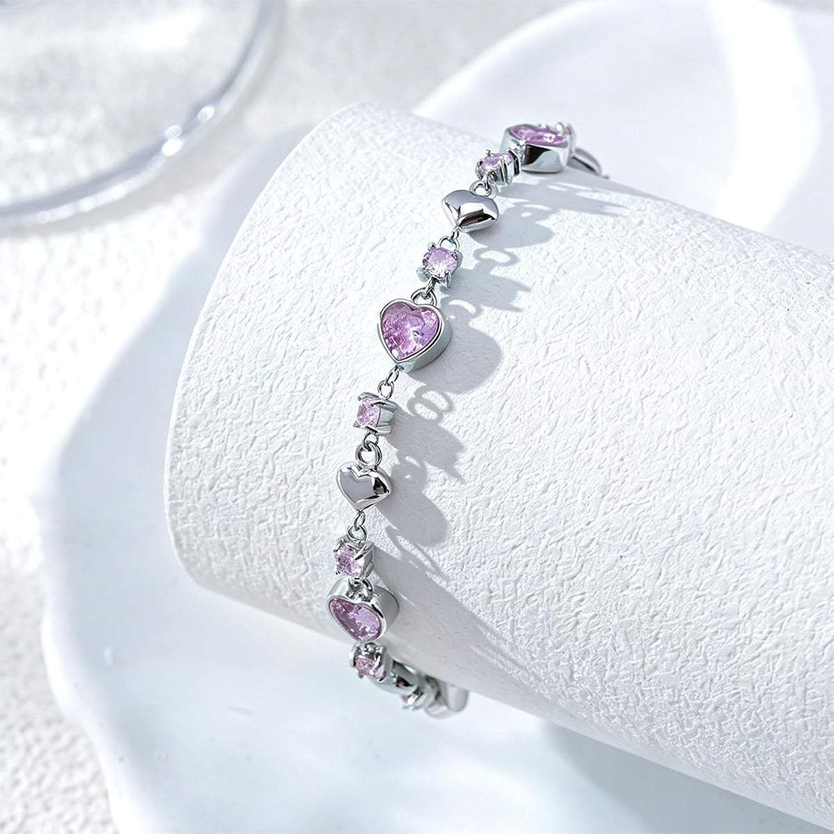A silver bracelet with alternating heart-shaped and round pink gemstones, displayed on a white textured cylindrical stand.