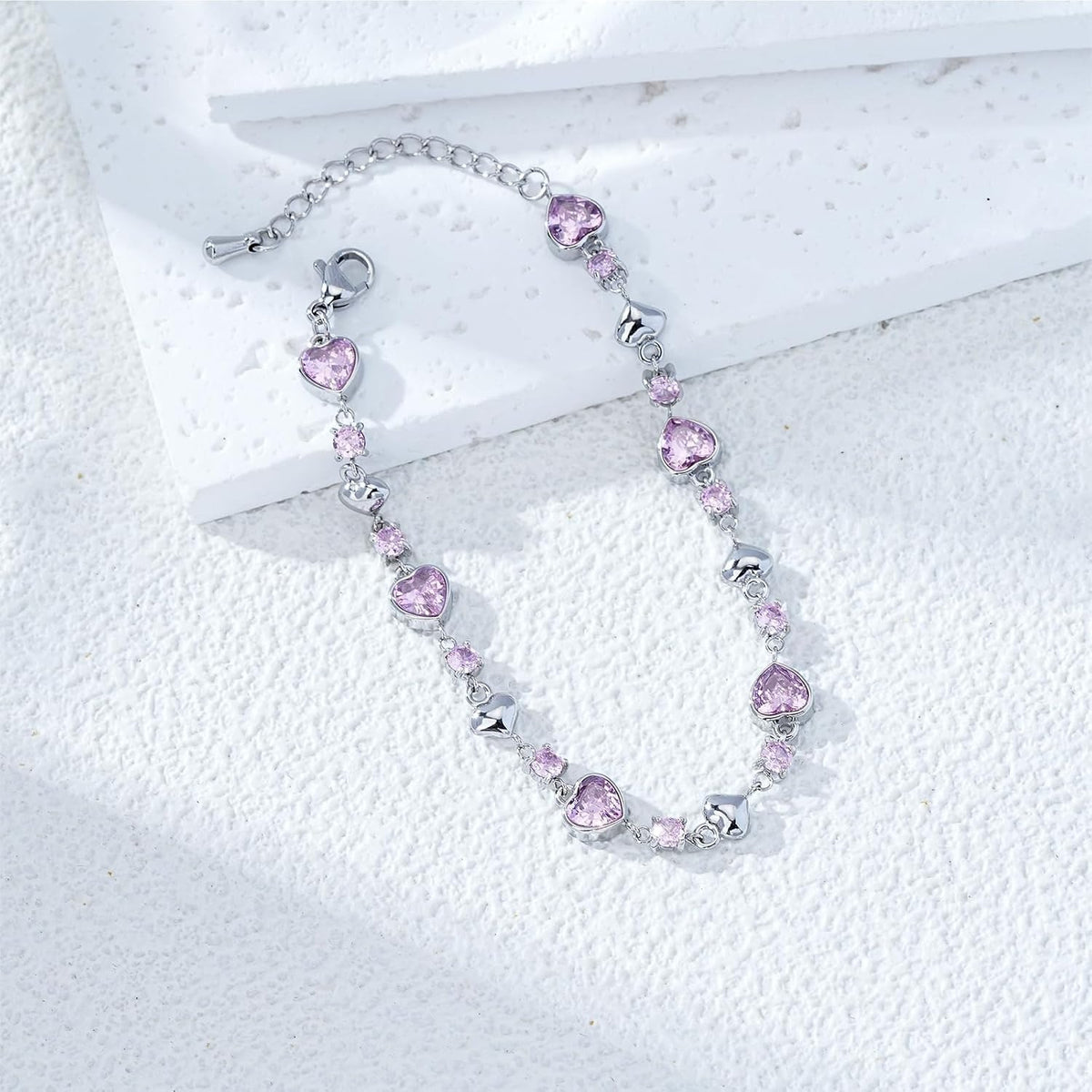 A silver chain bracelet featuring heart-shaped pink gemstones and small clear rhinestone accents, displayed on a textured white surface.