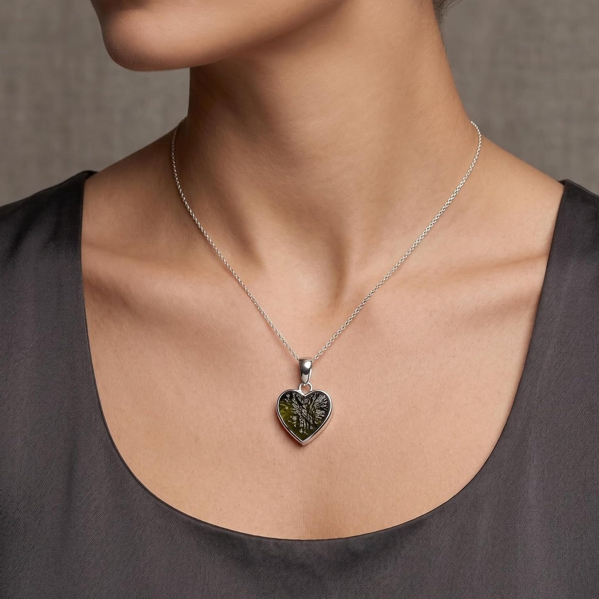 A close-up of a person wearing a delicate silver chain necklace with a heart-shaped pendant. The pendant features a dark green textured center with a silver tree design, worn over a dark gray scoop-neck top against a neutral gray background.