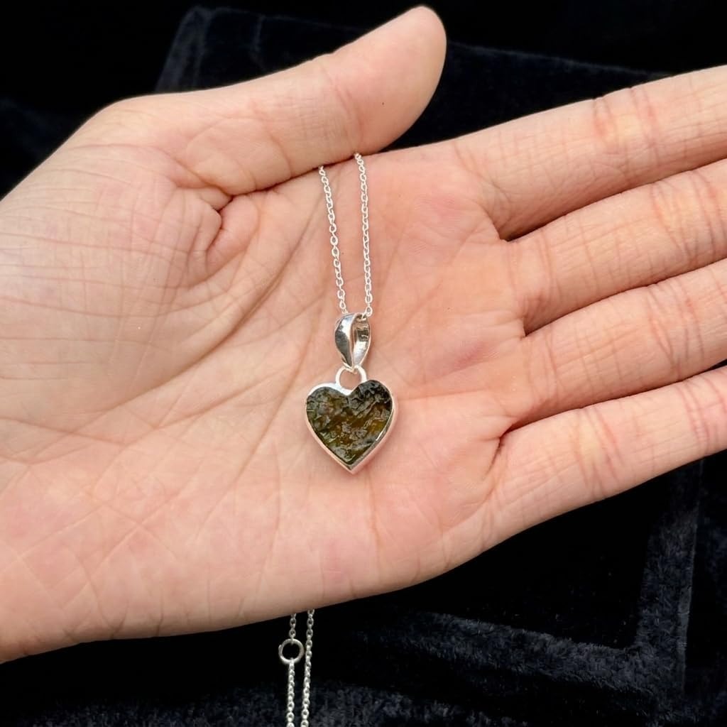 A heart-shaped pendant necklace with a green and gold patterned stone in a silver bezel, held in a person's open hand against a black velvet background.