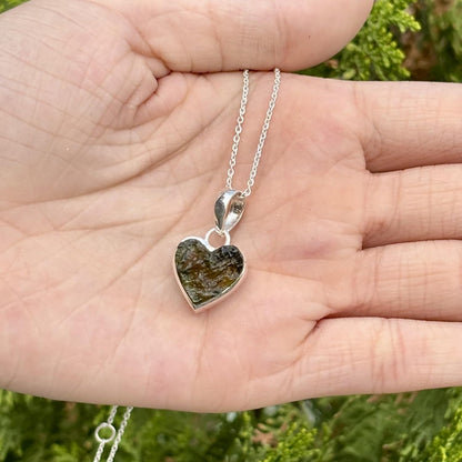A heart-shaped pendant necklace with a polished silver border and a textured green stone center, held in an open hand against a blurred green outdoor background.