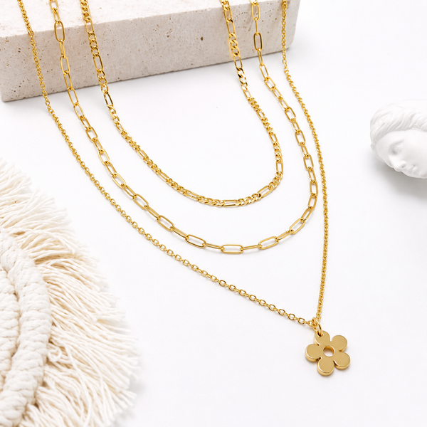 A gold layered necklace set displayed on a white surface with neutral props. The set includes a delicate chain, a paperclip chain, and a longer chain ending in a small flower-shaped pendant.