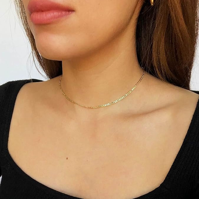 A close-up of a woman's neck and upper chest wearing a delicate gold chain necklace with a small central bead, paired with a black top.