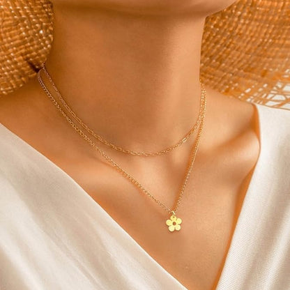 A close-up of a model wearing a delicate double-layered gold necklace with a small flower-shaped pendant, styled with a white top and a woven straw hat.