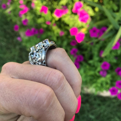 A close-up of a hand wearing a silver-toned ring with a large, ornate floral or scrollwork design, set against a blurred background of green foliage and pink flowers.