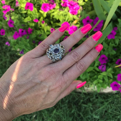 A silver filigree ring with a dark oval center stone is shown on a person's hand, with bright pink nails and a blurred background of green foliage and magenta flowers.